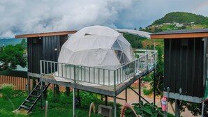 Une structure en forme de dôme géodésique, posée sur une plateforme dotée d’une rambarde métallique, entourée de verdure et avec un paysage vallonné en arrière-plan.