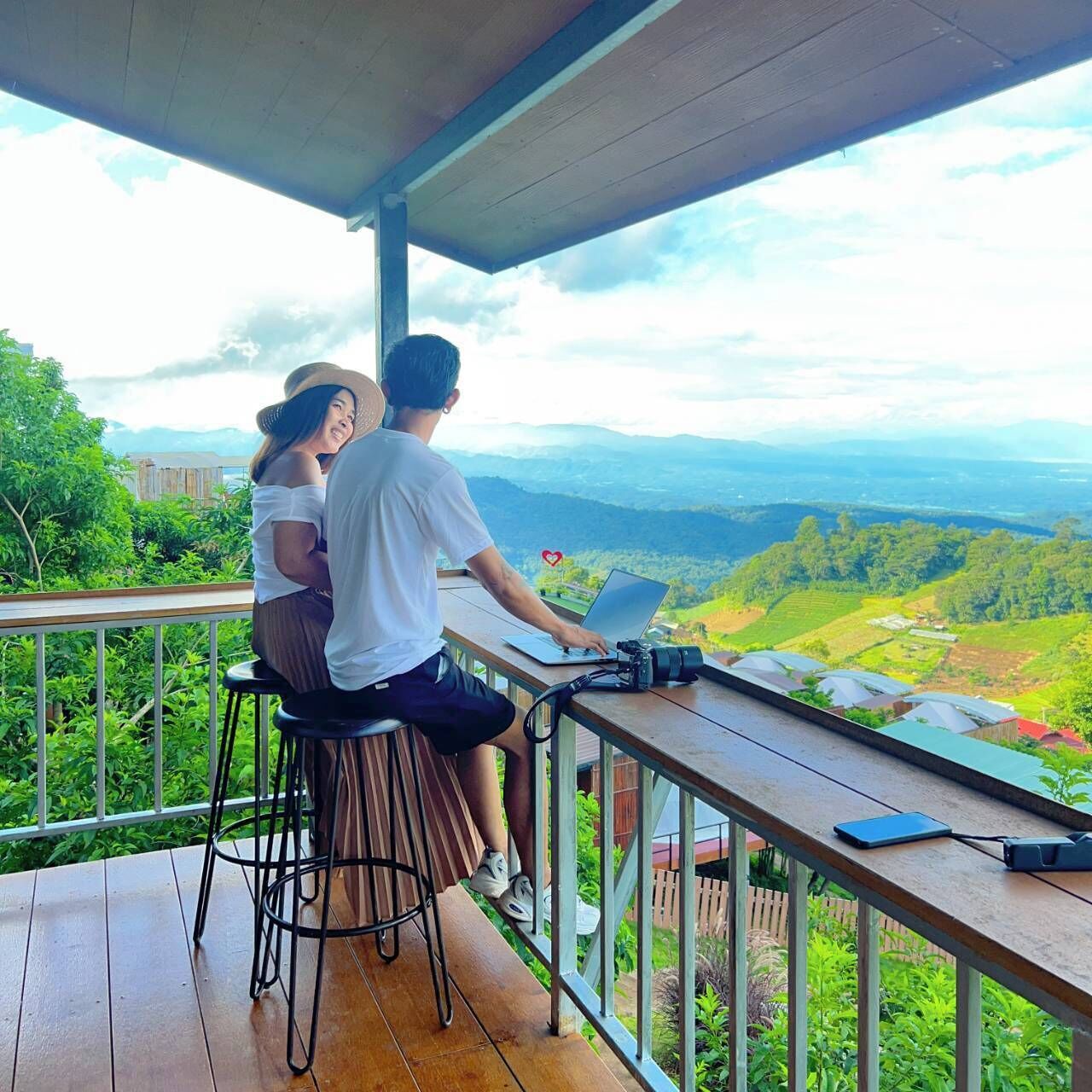 Una pareja sentada en una mesa de madera en un balcón con vistas a un paisaje montañoso.