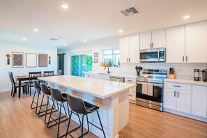 Une cuisine moderne avec des meubles blancs, un îlot central doté de tabourets, des appareils en acier inoxydable et une vue sur un espace extérieur verdoyant.