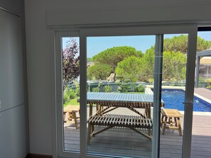 Blick durch eine Glastür auf einen Poolbereich mit Gartenmöbeln aus Holz und einem klaren, blauen Himmel.