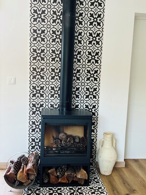 Ein Kaminofen mit schwarzem Schornstein, eine Wand mit geometrischem Muster und eine Vase auf dem Boden.
