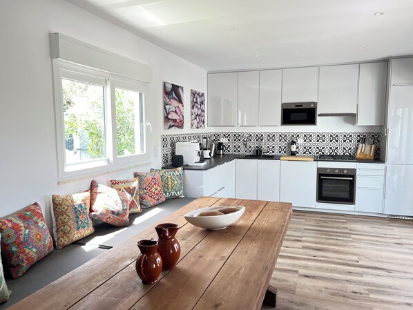 Een moderne keuken met witte kastjes, een houten bank met kleurrijke kussens en een groot raam.