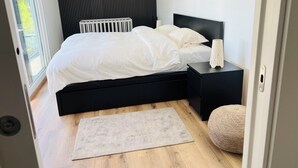 A modern bedroom with a black headboard, a white bedspread, a bedside table with a lamp, and a view of greenery through a window. - Sun, sea, and Lisbon—your vacation home is waiting for you (Costa de Caparica)