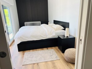 Ein modernes Schlafzimmer mit schwarzem Kopfteil, weißer Bettdecke, Nachttisch mit Lampe und Blick auf Grün durch ein Fenster.