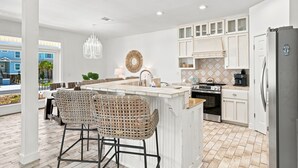 A modern kitchen with white cabinetry, a central island with bar stools, a hanging light fixture, and a view of the outside. - 🏄🌴Golf Cart | Steps to Beach | Multiple Balconies | Pelican Pod☀️ (Navarre)