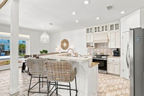 A modern kitchen with white cabinetry, a central island with bar stools, a hanging light fixture, and a view of the outside.
