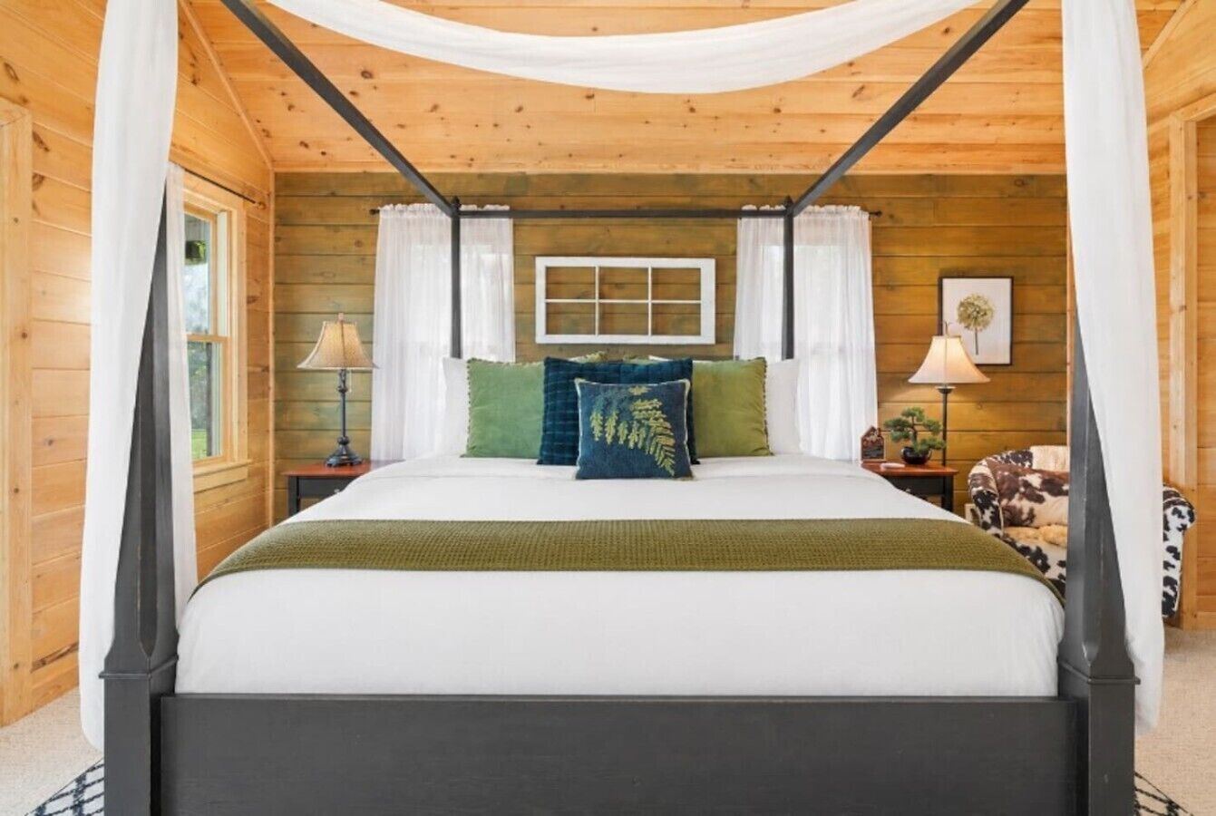A four-poster canopy bed with a white and green bedspread, two nightstands with lamps, and a cow-print chair.