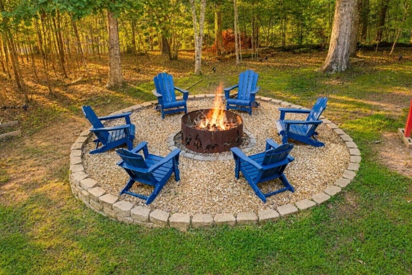 Una chimenea circular con sillas a su alrededor, ubicada en un área cubierta de césped con árboles al fondo.