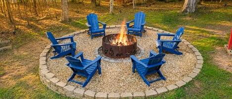 Una chimenea circular con sillas a su alrededor, ubicada en un área cubierta de césped con árboles al fondo.