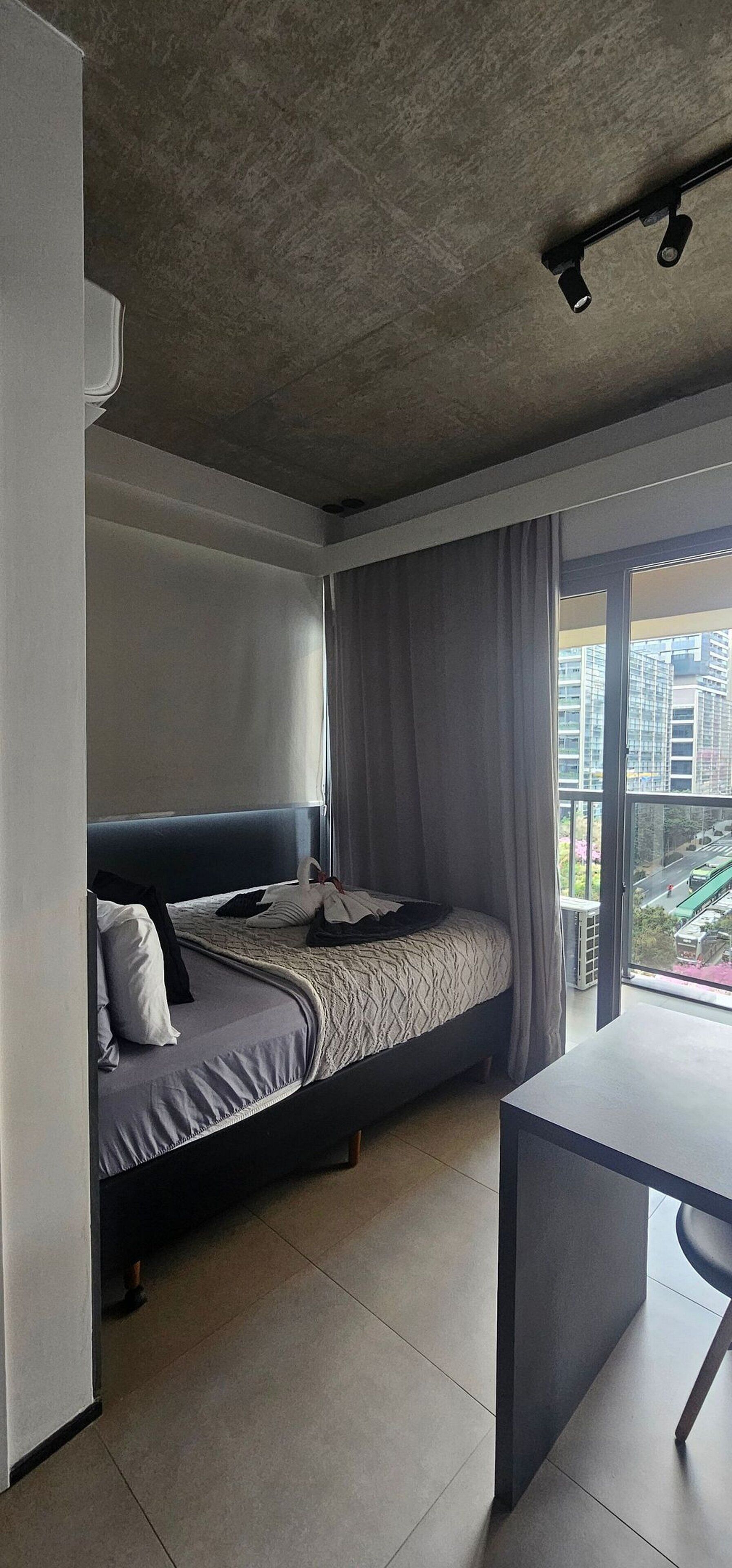 Quarto de hotel moderno com cama, escrivaninha e uma janela grande com vista para a cidade.