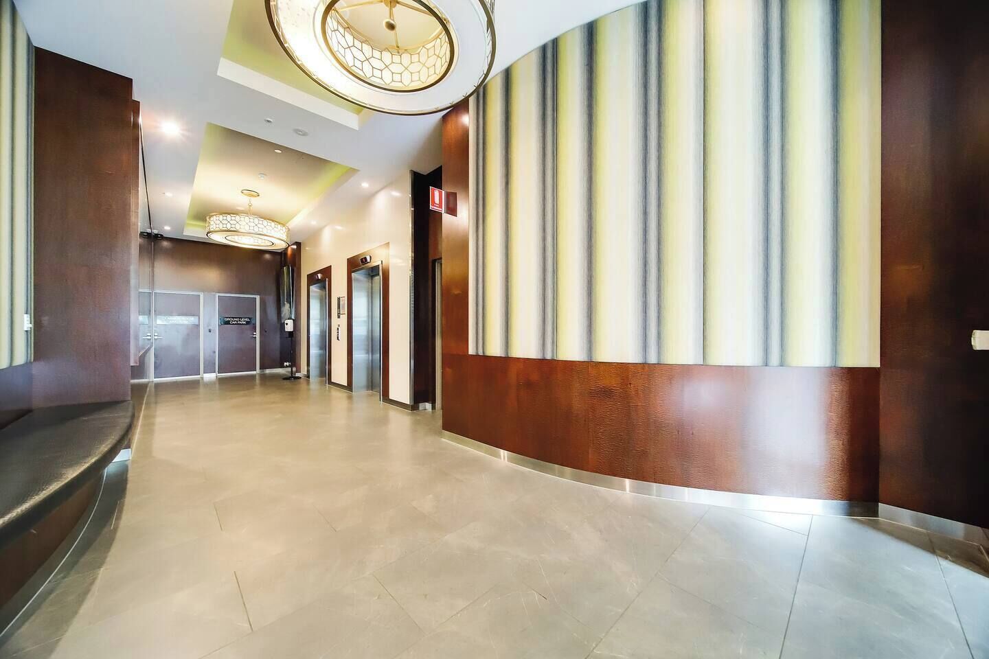 A hotel corridor with a modern design, featuring a central light fixture, a patterned wall, and a series of doors leading to rooms.