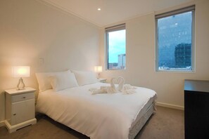 A hotel room with a large bed, two bedside tables, a dresser, and a window with a city view. - Amyra Residences on Rowlands Place - Unit 605 (Adelaide)