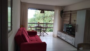 Uma sala de estar com um sofá vermelho, uma mesa de jantar com cadeiras, uma televisão e uma porta de vidro deslizante que dá para uma varanda.