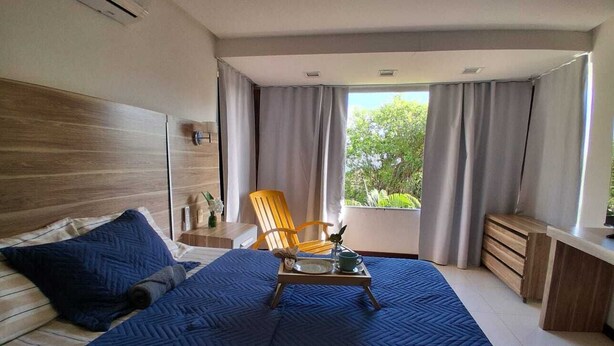 Quarto com uma cama grande, cabeceira de madeira, uma cadeira, mesa de cabeceira e janela com cortinas.