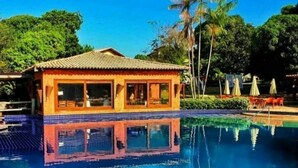 Um edifício junto à piscina, com telhado e janelas revestidas de azulejos, rodeado por uma piscina azul límpida que reflete o céu.