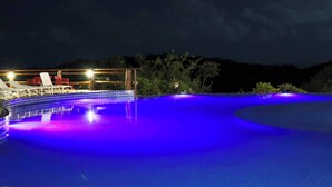 A swimming pool with a curved edge, surrounded by lounge chairs and a covered area with lights.