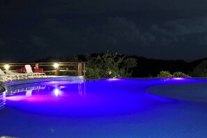 A swimming pool with a curved edge, surrounded by lounge chairs and a covered area with lights.