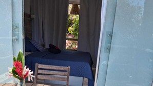 A wooden table with a vase of flowers, a chair, and a bed with a blue bedspread and white curtains.