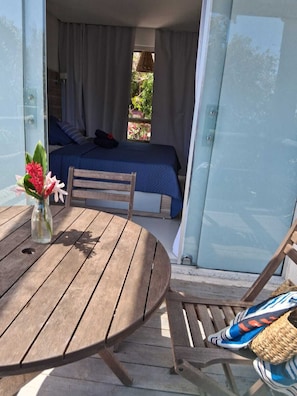 A wooden table with a vase of flowers, a chair, and a bed with a blue bedspread and white curtains.