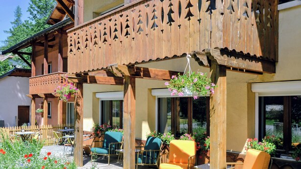 A wooden balcony with a decorative railing, outdoor seating with chairs and tables, and hanging flower baskets. - ABRIES HOTEL  (Abriès-Ristolas)