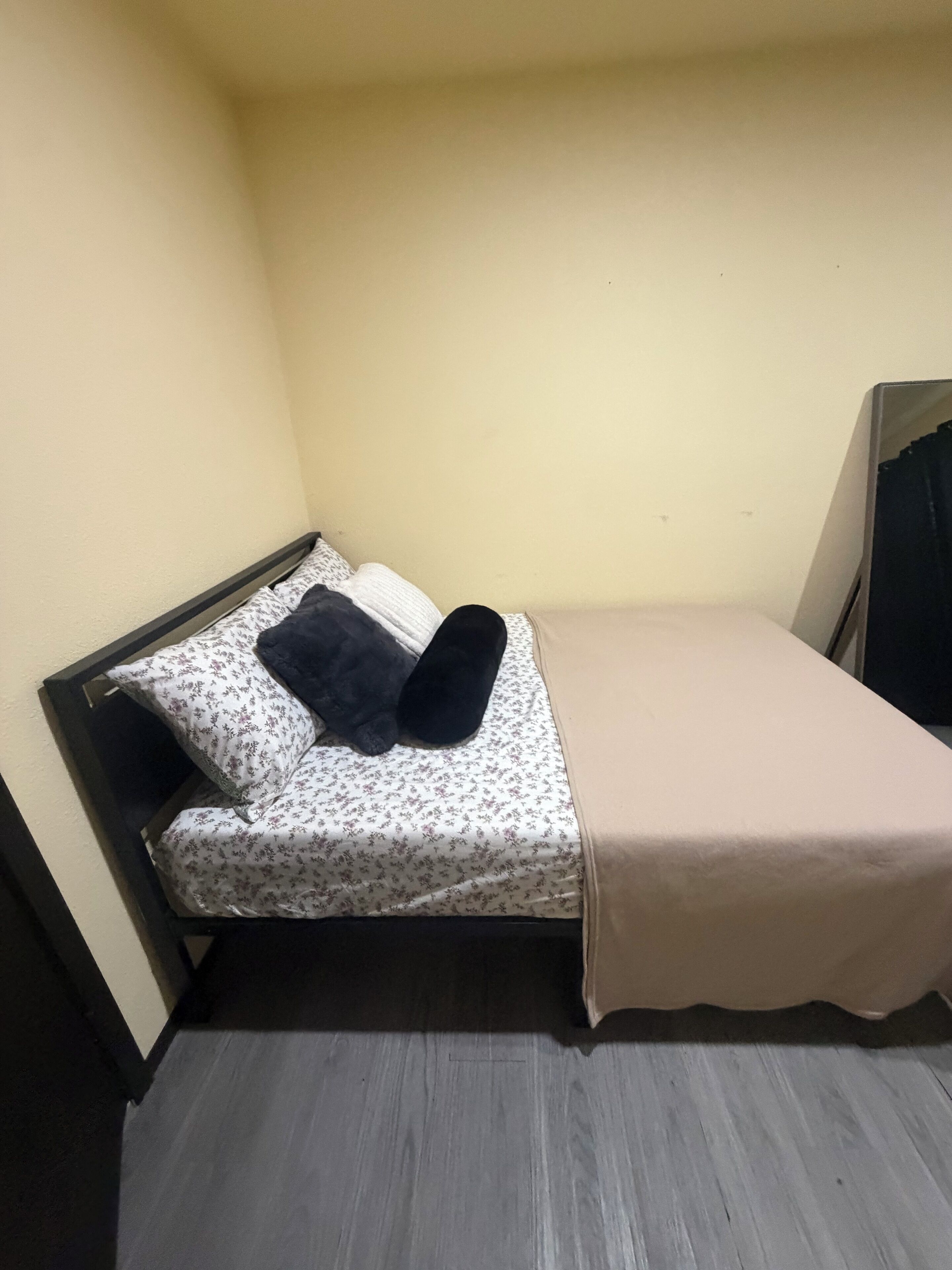 A small, single-bed room with a plain wall, a wooden floor, and a simple bed with a floral-patterned bedspread and two pillows.