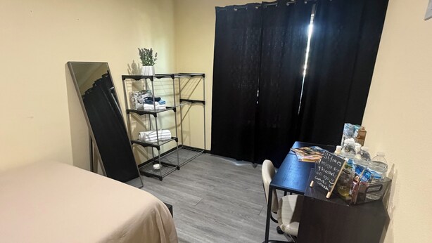 A room with a bed, a desk with a chair, a small shelf with items, and a black curtain.