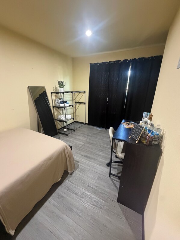A room with a bed, a desk with a chair, a small shelf with items, and a black curtain.