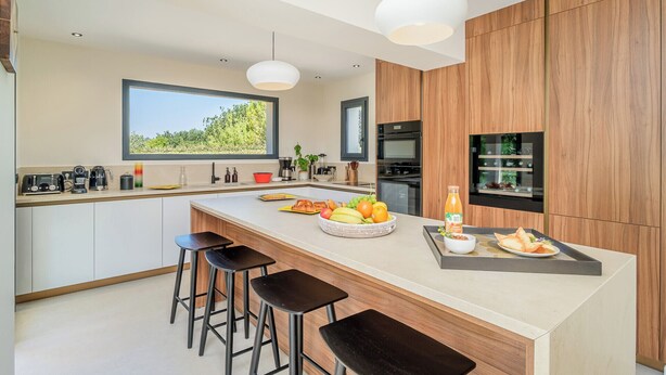 Een moderne keuken met een centraal eiland, barkrukken, een ingebouwde oven en uitzicht op groen.