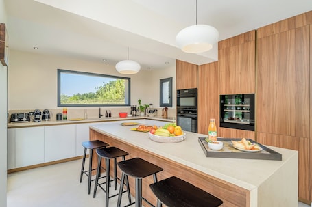 A modern kitchen with a central island, bar stools, a built-in oven, and a view of greenery outside.