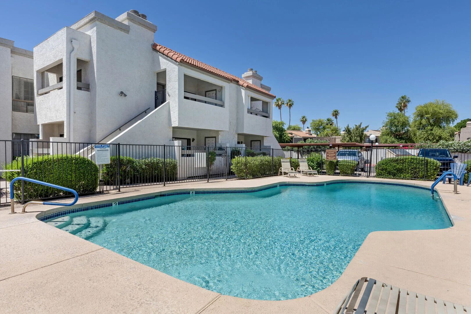 A swimming pool with a curved edge, surrounded by a fence, and adjacent to a multi-story building with balconies.