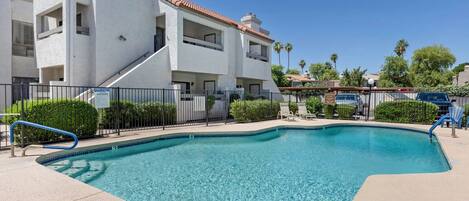 A swimming pool with a curved edge, surrounded by a fence, and adjacent to a multi-story building with balconies.