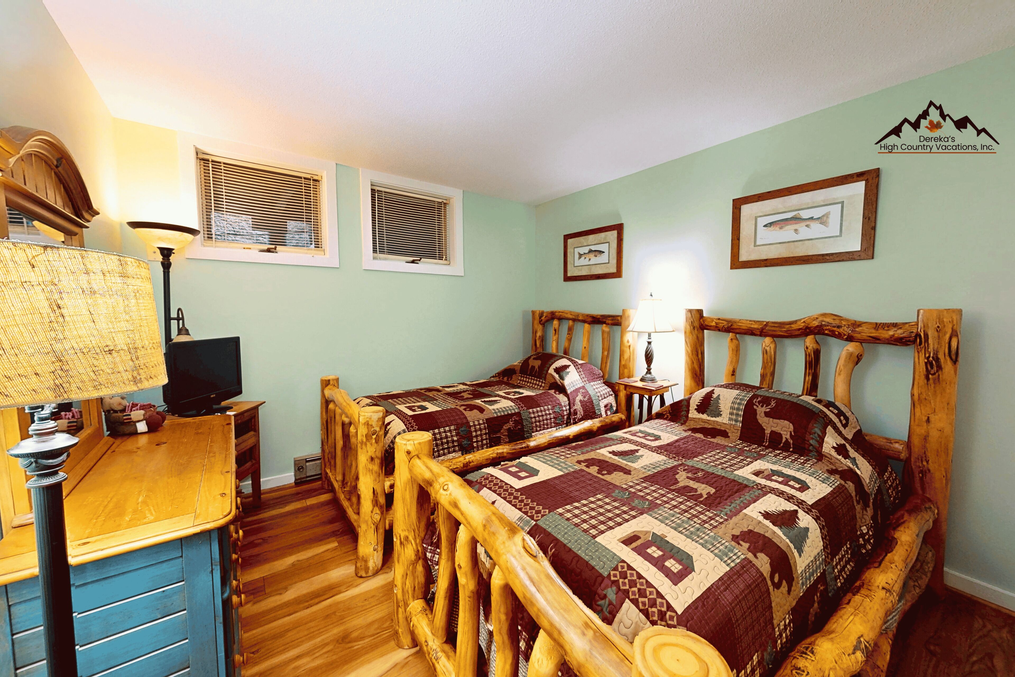 Ein Zimmer mit zwei Holzbetten, einem Schreibtisch mit Lampe und einem Fernseher. Zwei Fenster mit Jalousien und ein Bild von Fischen im Bilderrahmen an der Wand.