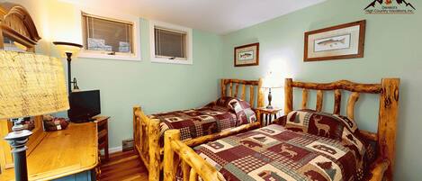 A room with two wooden beds, a desk with a lamp, and a television. There are two windows with blinds, and a framed fish picture on the wall.