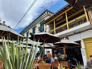 An outdoor seating area with tables and umbrellas, a building with a balcony, and a clear sky. - El Retiro apartment – peaceful, quiet, yet close to downtown with LOVELY views! (Retiro)