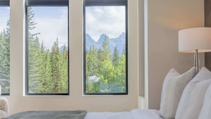 Habitación de hotel con cama, lámpara y vistas a un bosque y montañas a través de amplias ventanas.