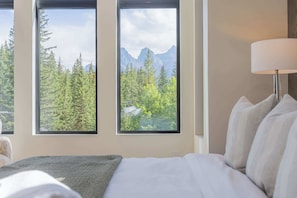 Ein Hotelzimmer mit einem Bett, einer Lampe und Blick auf einen Wald und Berge durch große Fenster.