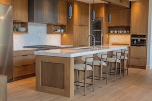 A modern kitchen with a central island, bar stools, wooden cabinetry, and built-in appliances. - Ultra-Luxury Estate w/ Nordic Spa & Concierge (Canmore)
