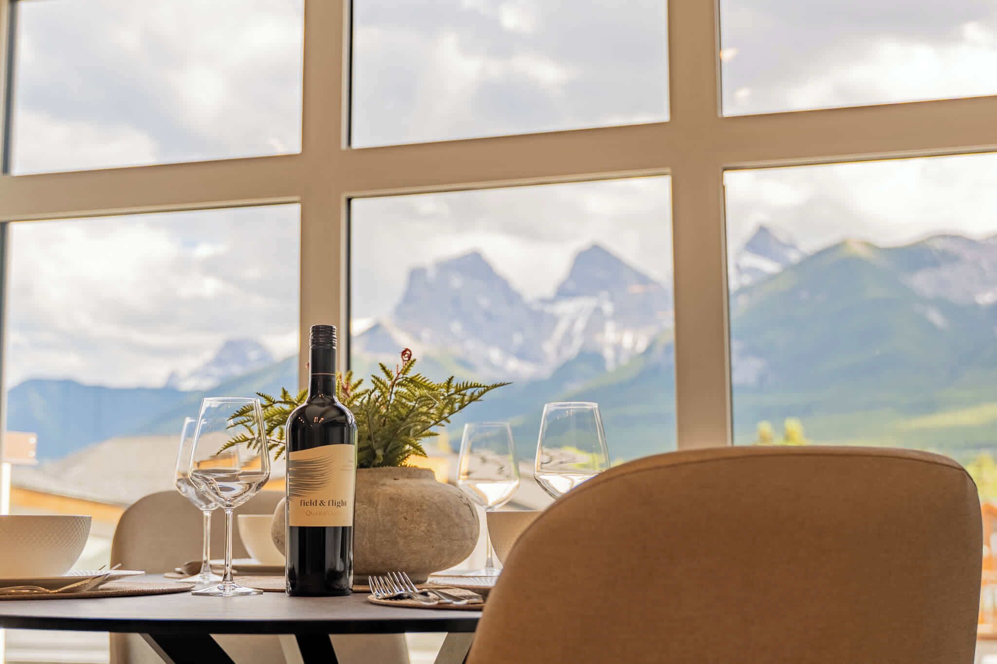 A dining table with a bottle of wine, glasses, and a plant, set against a backdrop of mountains through large windows.
