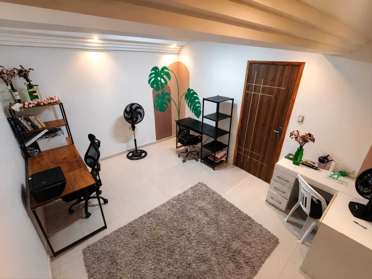 A room with a desk, chair, bookshelf, two plants, a fan, and a door.