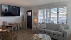 En stue med en grå sofa, et glasbord, et fjernsyn og et vindue med persienner.