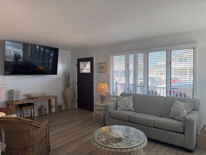 Ein Wohnzimmer mit grauem Sofa, einem Glastisch, einem Fernseher und einem Fenster mit Jalousien.