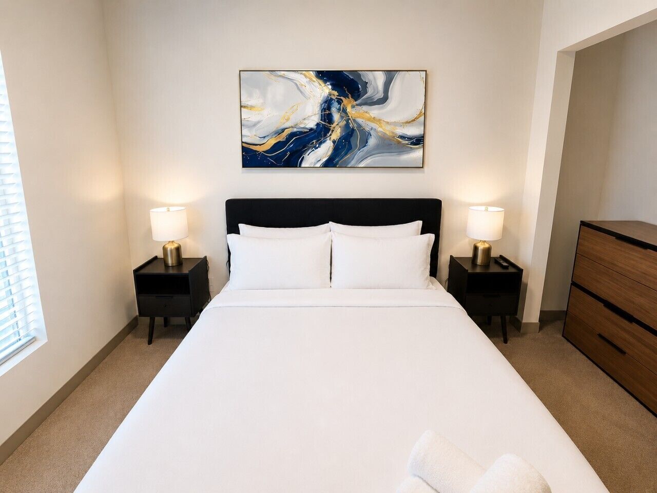 Une chambre d’hôtel avec un grand lit, deux tables de chevet avec des lampes, une commode en bois et un tableau abstrait accroché au mur.