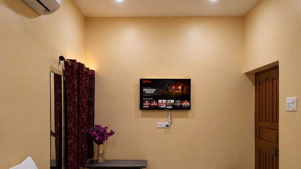 Quarto de hotel com cama, mesa de cabeceira com vaso, televisão na parede exibindo opções de filmes e porta de madeira.