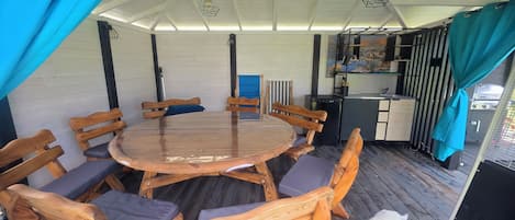 A covered outdoor dining area with a round wooden table and chairs, a grill, and a kitchen area.