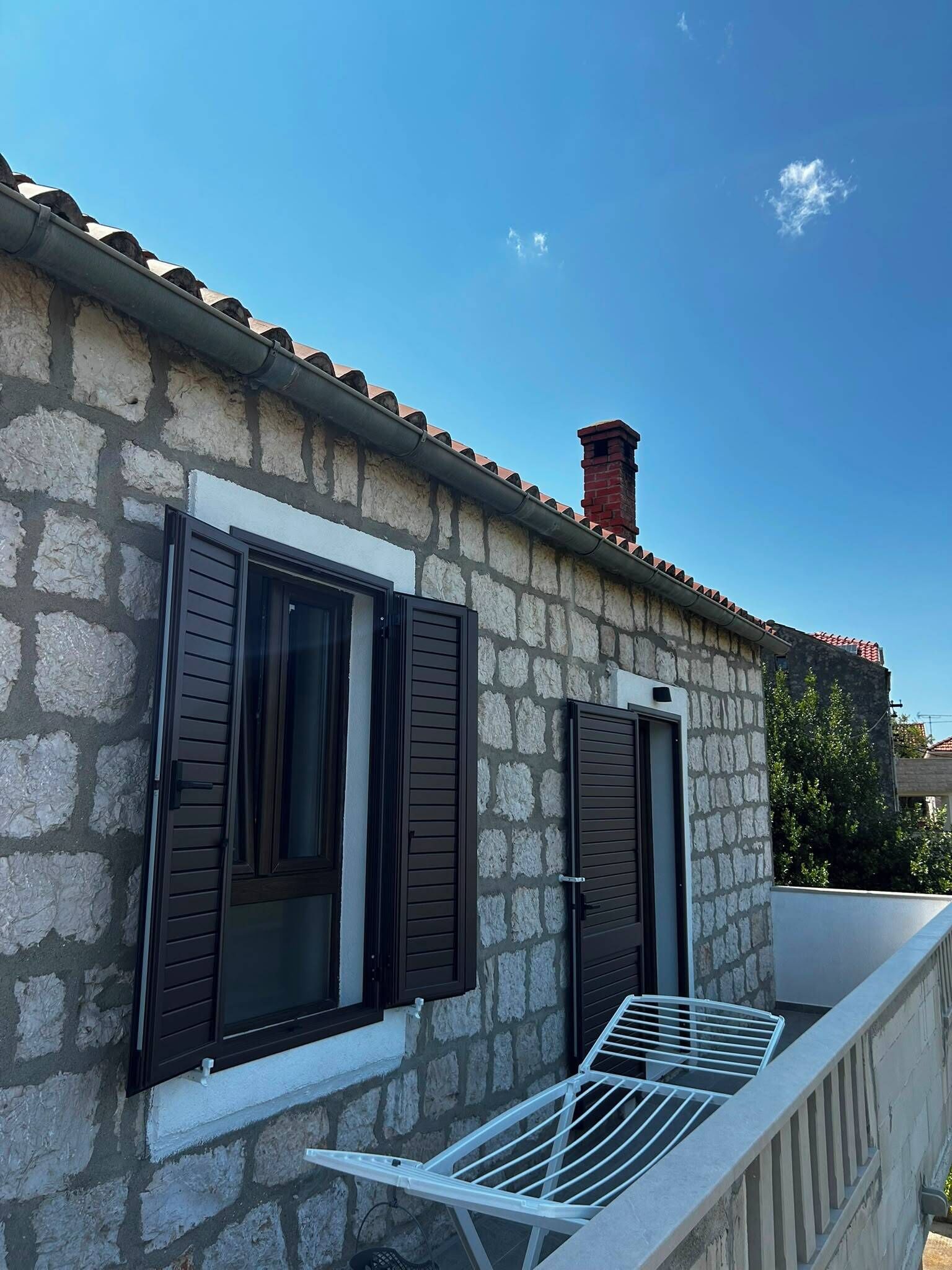 A stone building with two windows, a balcony with a metal railing, and a chimney.