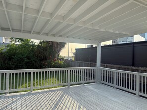 Eine überdachte Terrasse im Freien mit weißer Reling, einer flachen Decke und Blick auf ein dunkles Gebäude sowie grüne Bepflanzung im Hintergrund.