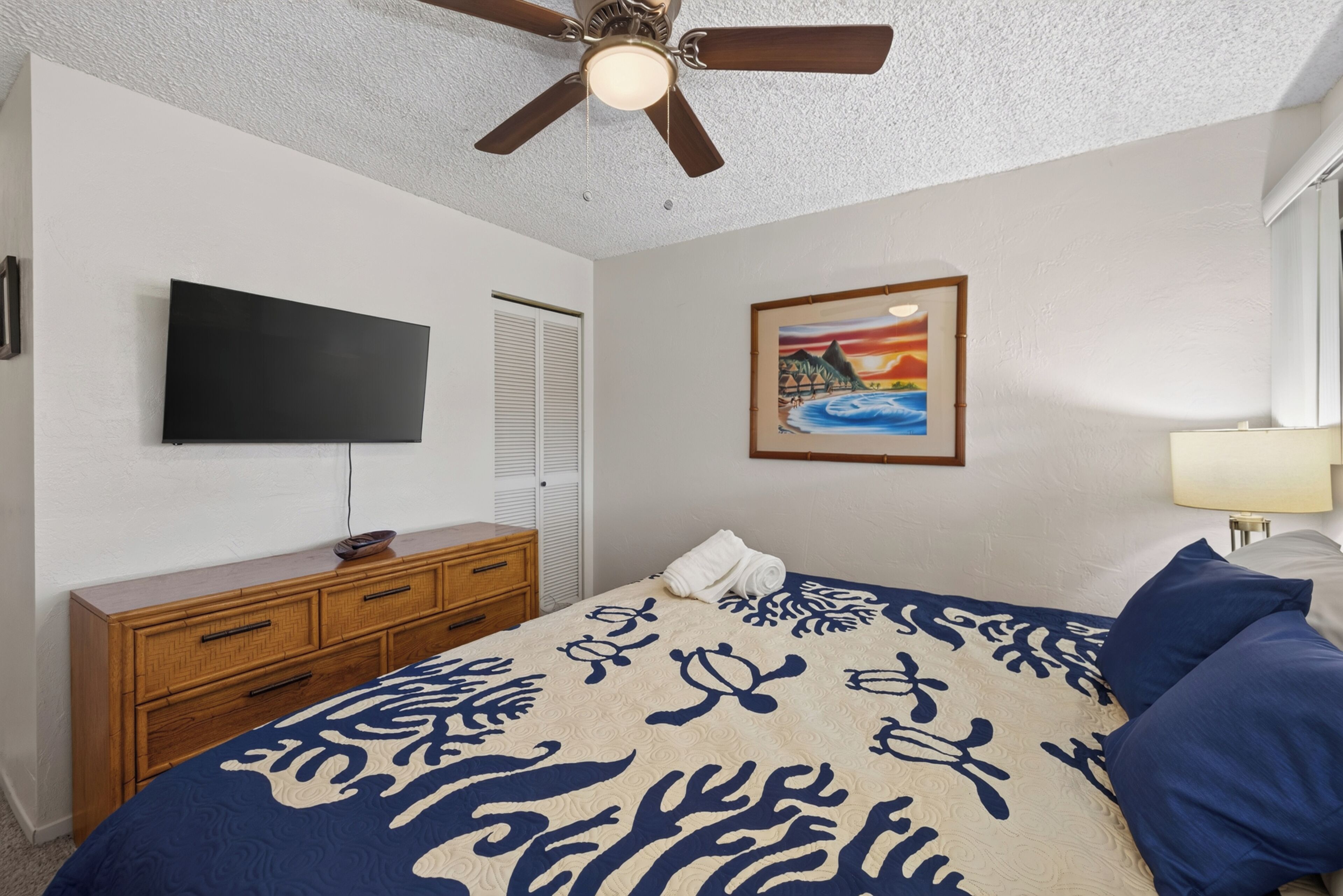 A bedroom with a bed, a television, a ceiling fan, a dresser, and a painting on the wall.