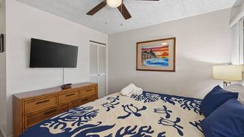 A bedroom with a bed, a television, a ceiling fan, a dresser, and a painting on the wall.