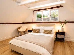 Ein Schlafzimmer mit schrägem Dach, einem großen Bett, einem Nachttisch mit Lampe, einem Sessel und einem Fenster mit Jalousien.