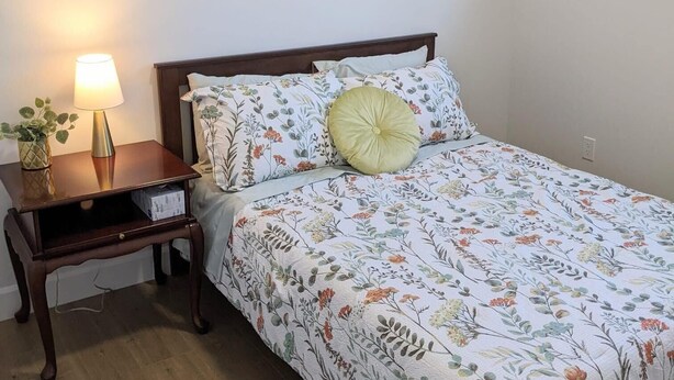 A bedroom with a bed, a nightstand, a lamp, a framed botanical illustration, and a window. - Steps to Beach • 2BR White Rock Suite • Sleeps 4 (Surrey)
